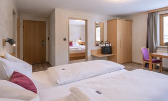 A modern hotel room with two beds and neutral colors. The room includes furniture such as a desk, a wardrobe, and a mirror.