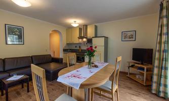 A bright and inviting apartment with a cozy seating area and a dining table. The kitchen is modernly equipped, and there are fresh flowers on the table.