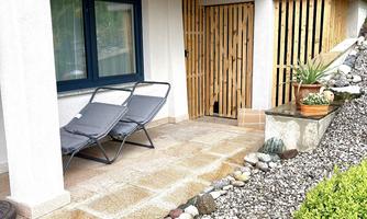 A cozy outdoor area with two loungers and a well-maintained stone path. Green plants and wooden cladding give the entrance an inviting character.
