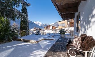 A wintry view of snow-covered mountains and a cozy terrace. The sky is clear and blue, creating a serene atmosphere.