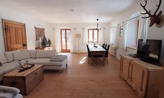 A cozy living space with bright walls and a wooden floor. The furnishings consist of a comfortable sofa, a dining table, and a television.