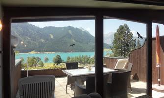 A beautiful view of a clear lake surrounded by green mountains. In the foreground, there is a terrace with cozy seating.
