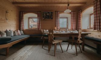 A cozy room with wooden walls and a large table in the center. Next to it is a bench with cushions and blackout curtains at the windows.