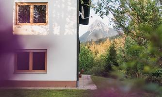A modern house with large windows stands in a green environment. In the background, an impressive mountain can be seen.