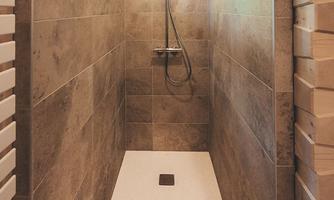 A modern shower with gray tiles and a large, white shower tray. The walls are made of wood and there are windows for daylight.