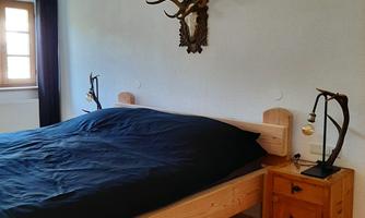 A cozy bedroom with a large bed and a dark blue duvet. On the wall, there are antlers and a lamp, and the floor is made of wood.