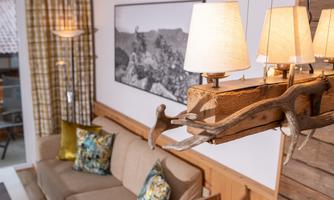 A cozy living room with a sofa and colorful cushions. A decorative lamp made of wood and antlers hangs above the room.