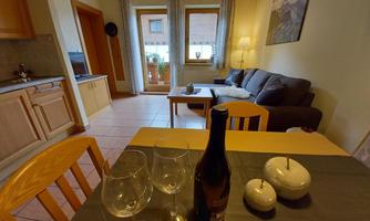 A cozy apartment with a dining table, glasses, and a bottle of wine. In the background, a kitchen and a living room with a couch can be seen.