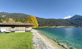 A calm lake with clear water and a green meadow. In the background, mountains and a wooden building can be seen.