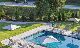 A clear pool with loungers and sun umbrellas in a well-maintained garden. In the background, two motorcycles can be seen on a road.