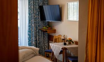 A cozy hotel room with a bed, a table, and two chairs. A television hangs on the wall, and there is a large window front with curtains.
