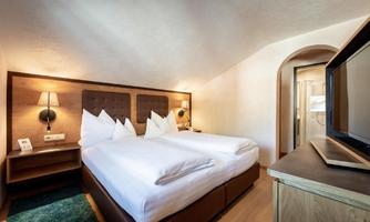 A modern hotel room with two beds, bright pillows, and elegant wooden furniture. The room is well-lit and offers a cozy living area.