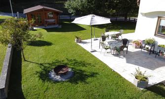 A beautiful garden with a terrace and a wooden house in the background. The garden has a well-kept lawn and a seating area with a sunshade.