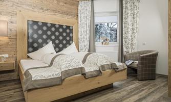 A modern bedroom with a large wooden bed and elegant bed linens. In the background, there are windows with curtains and a cozy armchair.