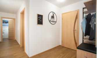 A bright hallway with wooden flooring and white walls. A decorative sign hangs on the wall, and to the right is a coat rack.
