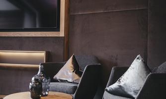 A cozy living area with two modern armchairs and a round table. In the background, there is a television and the walls are designed in warm tones.