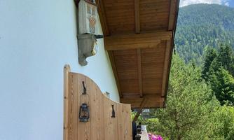 A cozy balcony with wooden cladding and blooming plants. In the background, green mountains and a blue sky can be seen.