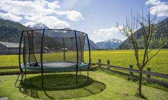 A trampoline stands on a green meadow with a view of the mountains. Behind the trampoline, a blooming yellow field stretches under a clear blue sky.