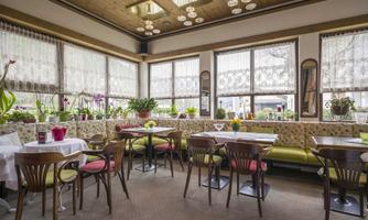 A cozy restaurant with tables and chairs.  
Large windows let in a lot of light and the room is decorated with plants.