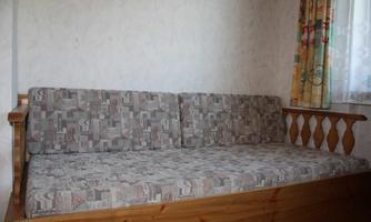 A cozy wooden couch with a gray patterned cushion cover. Next to the couch is a window with colorful curtains.