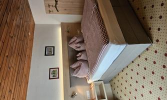 A cozy bedroom with a large bed and checkered pillows. The walls are bright, and the floor has a beautiful carpet with a floral pattern.