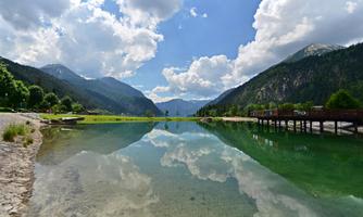 A clear lake surrounded by mountains and lush greenery. The sky is blue with some white clouds reflecting in the water.