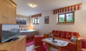 A cozy living area with a red sofa and a television. In the background, there is a bedroom with a double bed and windows that let in natural light.