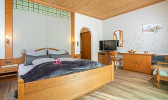 A cozy room with a double bed, wooden details, and a flat-screen TV.  
There is a desk and a chair as well as a decorative wall with soft colors.