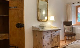 A cozy bedroom with wooden flooring and a large window. An adjoining room with an antique wardrobe, a mirror, and a lamp creates an inviting atmosphere.
