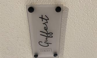 A transparent sign with the inscription "Guffert". It is attached to a white wall and secured with black brackets.