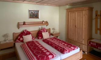 A cozy bedroom with a large double bed and red bed covers. The room is bright and lovingly decorated, with wooden furniture and a decorative dresser.