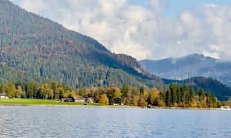A tranquil landscape with a lake and gentle hills in the background. The trees display autumn colors under a clear sky.
