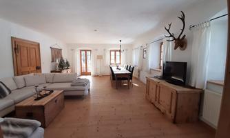 A modern living room with a cozy sofa and a wooden table. The walls are bright, and there are large windows that let in plenty of daylight.