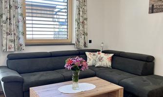 A cozy living room with a dark couch and a wooden table. The room has light curtains and a large window that lets in daylight.