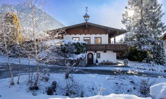 A picturesque house in the snow, surrounded by snow-covered trees. The sun is shining and provides a cozy winter atmosphere.