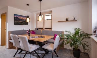A cozy dining area with a wooden table and chairs. On one wall, there is a plant and a picture, while the room lighting is accented by two pendant lights.