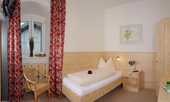 A cozy single room with wooden paneling and a window. There is a chair, a nightstand with a lamp, and fresh flowers.