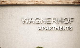 A modern sign reading "Wagnerhof Apartments" in front of a wood exterior. Colorful flowers and plants adorn the area around the sign.