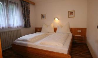 A simple bedroom with a double bed and two white pillows. The walls are light and there is a window with curtains.
