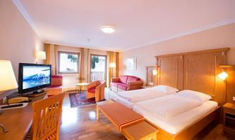 A bright hotel room with wooden interior and two single beds. There is a TV, seating, and large windows that let in plenty of light.