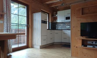 A modern kitchen in a wooden house with large windows. The room has light walls and a warm wooden floor.