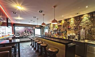 A modern bar interior design with a long counter and stylish chairs. The walls are adorned with soft lighting elements and a decorative stone wall.
