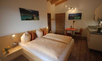 A cozy bedroom with a double bed and wooden details. The room is bright and welcoming, with a small dining table and artworks on the walls.