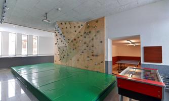 A climbing wall with colorful holds and a soft mat in the foreground. On the right, a tabletop game can be seen.