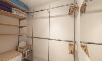 A bright room with open shelves and a clothing rod. On the left side are some cushions and a small wooden bench.