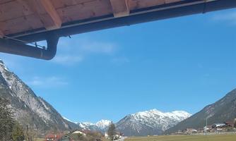 A beautiful mountain landscape with snow-covered peaks and clear blue skies. In the foreground, a meadow and a parking lot are visible.