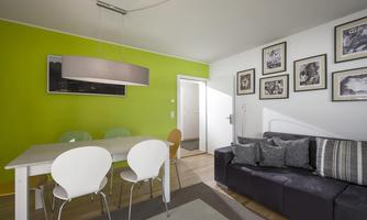 A modern living room with a green wall and light furniture. There is a dining table with chairs and a comfortable sofa.