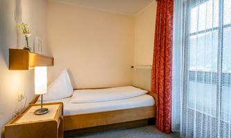A simple bedroom with a single bed and a lamp. The room has large windows with curtains and a bright, minimalist style.