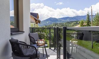 A cozy balcony with two chairs and a small table. In the background, green mountains and a blue sky can be seen.
