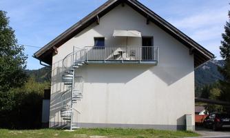 A simple house with a balcony and a metal spiral staircase. In the background, trees and mountains can be seen.